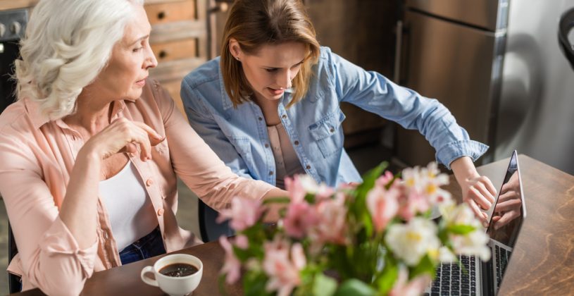 Senior woman and adult daughter at a kitchen table with coffee creating a moving plan on a laptop.