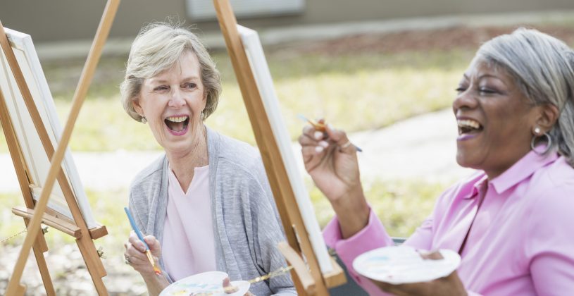 Two senior artists painting outdoors – both women are holding palettes and are sharing a laugh as they paint at easels. Finding opportunities to enjoy creative pursuits has been shown to increase happiness