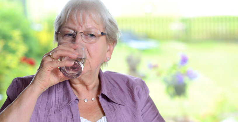 A senior woman wearing a crushed silk lavender top drinks a glass of water. Dehydration can can occur for many reasons besides increased heat exposure