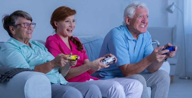 Seniors sitting on a couch with a caregiver playing a video game. Recent research shows a direct positive correlation between video game playing and cognitive abilities